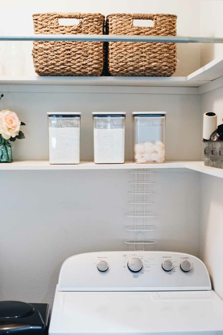 Small laundry closet with white shelves, baskets, and clear detergent containers.