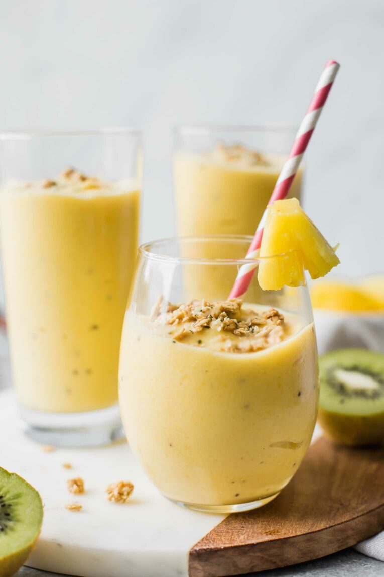 Yellow fruit smoothie on a marble cutting board with a straw and pineapple garnish.
