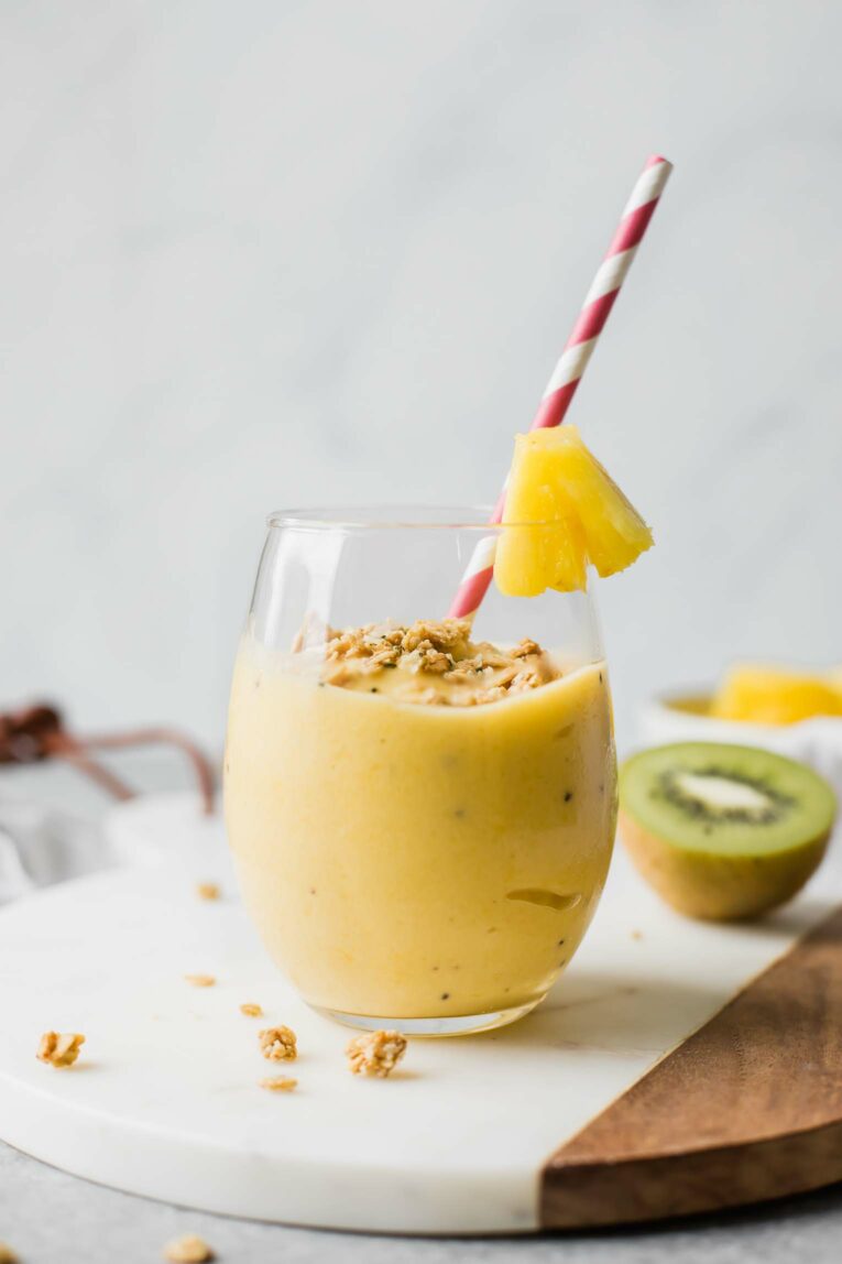 Yellow tropical smoothie in a glass cup with a straw and kiwi in the background.