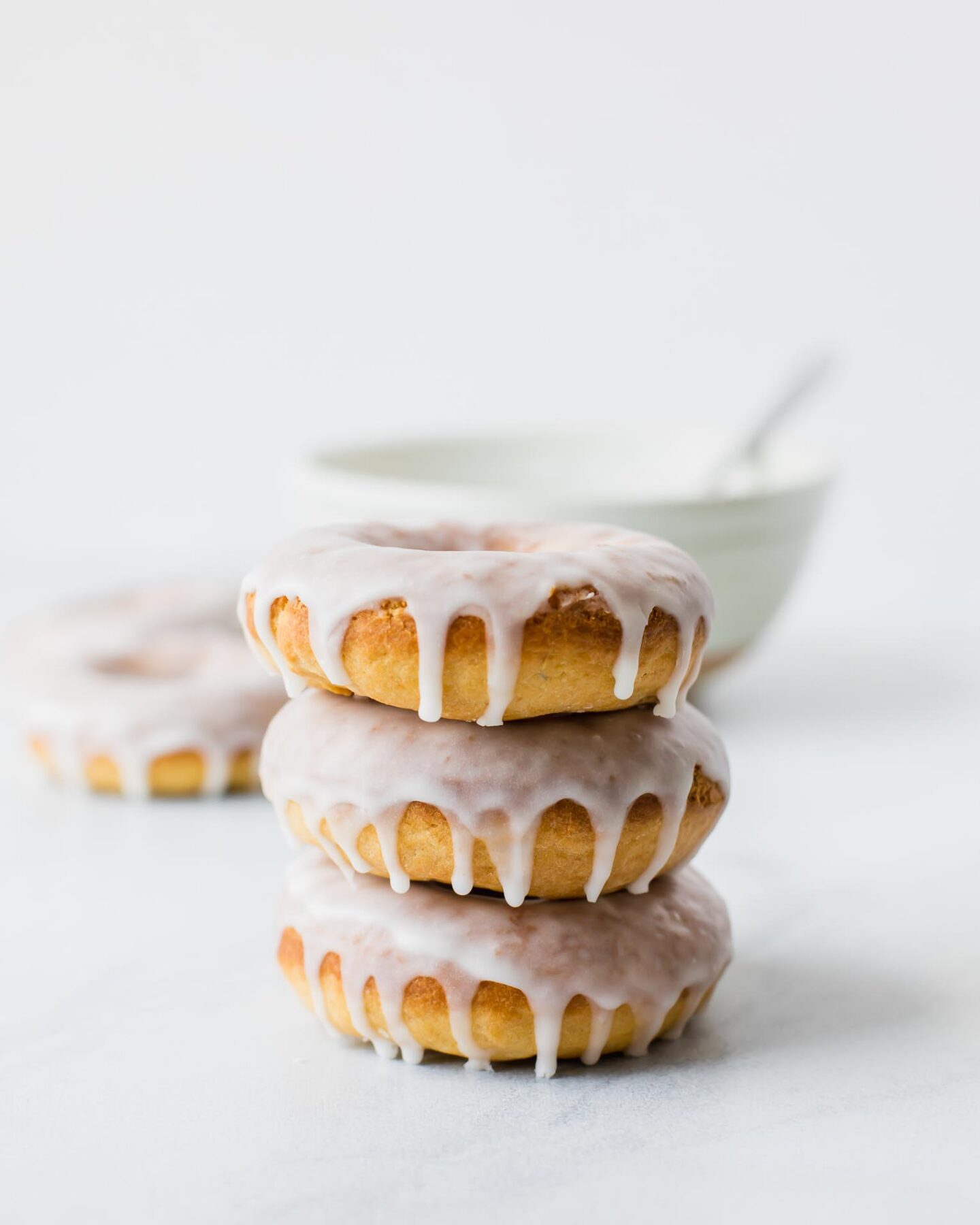 Stack of baked glazed doughnuts with a white bowl in the background.