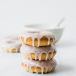 Stack of baked glazed doughnuts with a white bowl in the background.