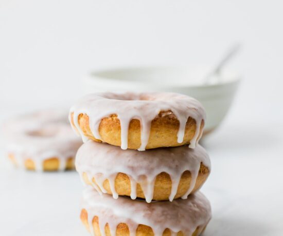 Stack of baked glazed doughnuts with a white bowl in the background.