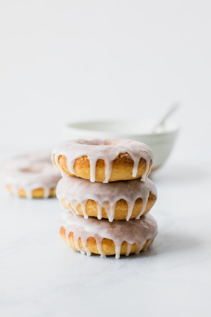 Stack of baked glazed doughnuts with a white bowl in the background.