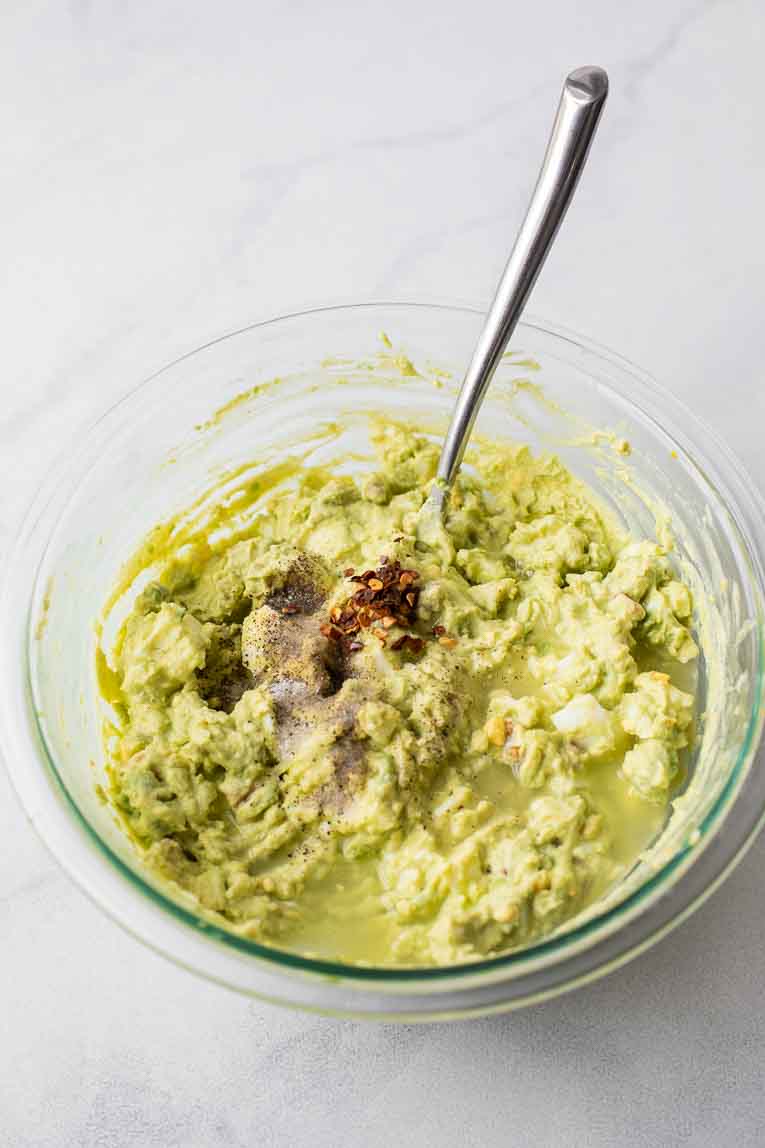 Glass bowl with mashed avocado, eggs, yogurt, walnuts, and crushed red pepper.