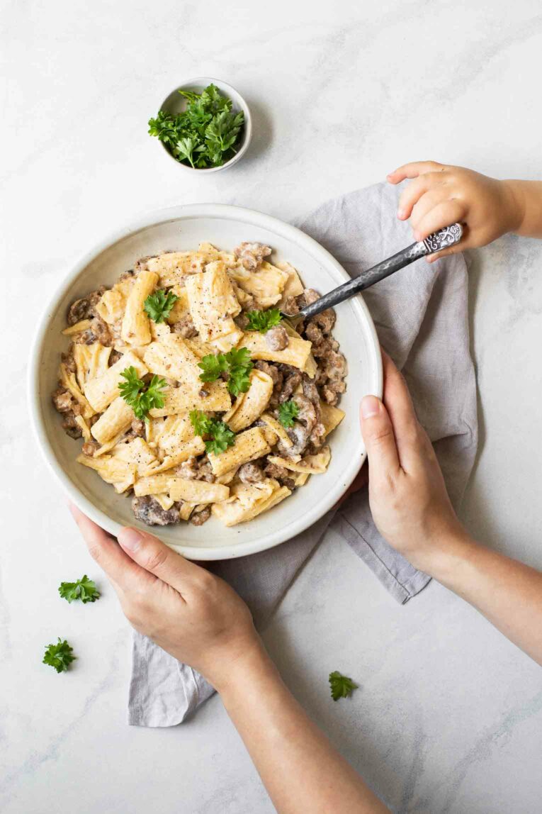 Hands holding a white bowl of Creamy Mushroom Sausage Rigatoni.