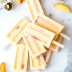 Top shot of Homemade Creamy Peach Popsicles, peach slices, and a napkin on a marble board.