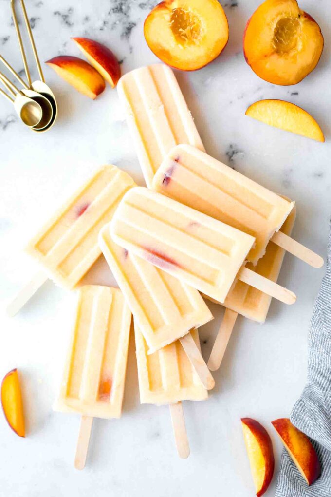 Top shot of Homemade Creamy Peach Popsicles, peach slices, and a napkin on a marble board.
