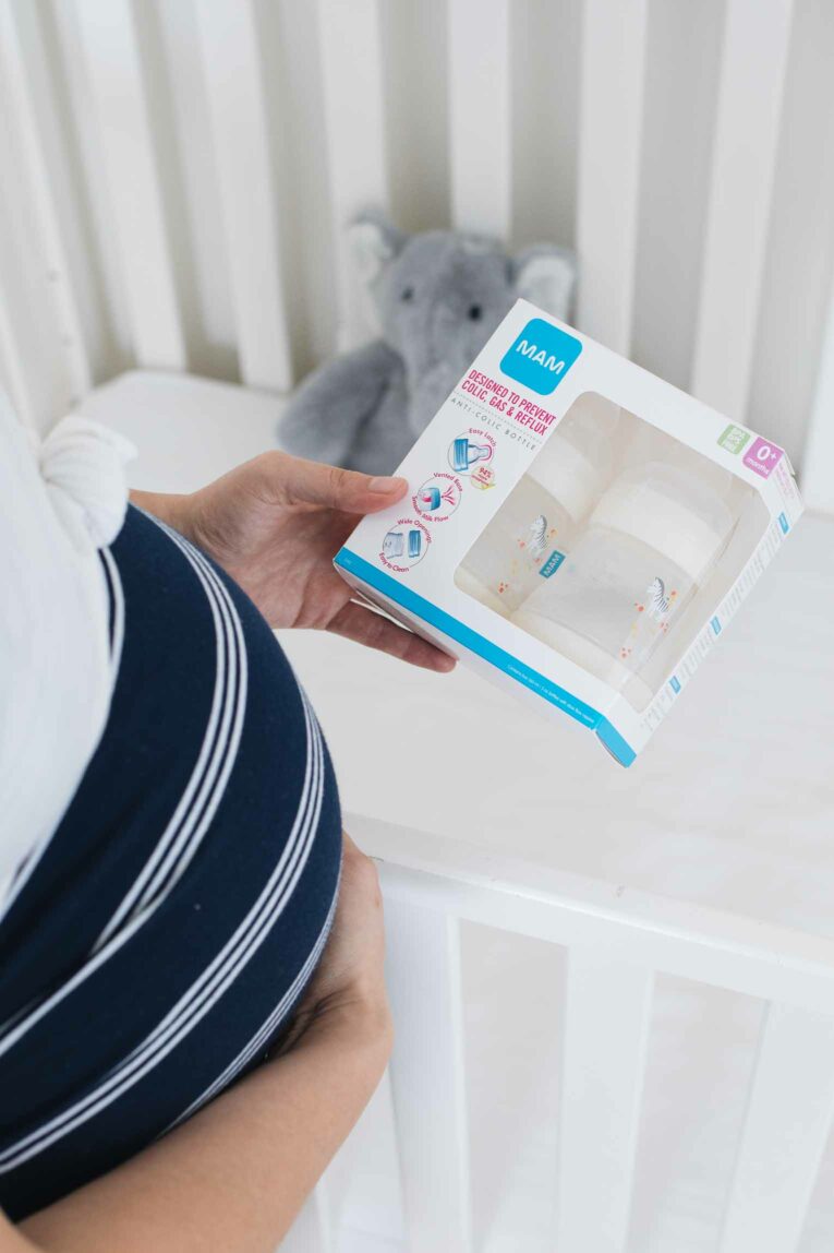 Pregnant mother holding MAM baby bottles over crib.