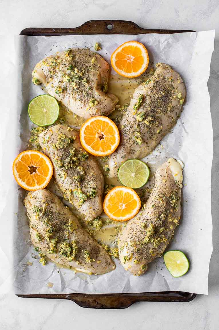 Raw marinated chicken, orange slices, and lime slices on a baking sheet.