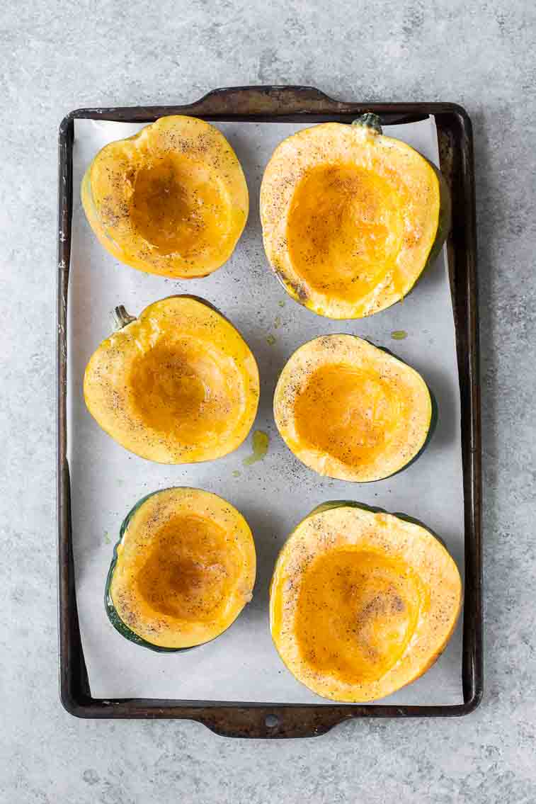 Raw acorn squash halved and drizzled with oil on a baking sheet.