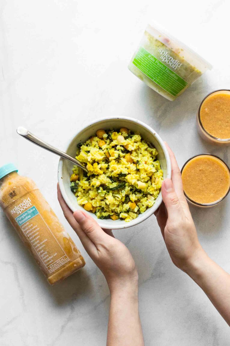 Two hands holding bowl of Splendid Spoon curry rice next to smoothies and drinks.