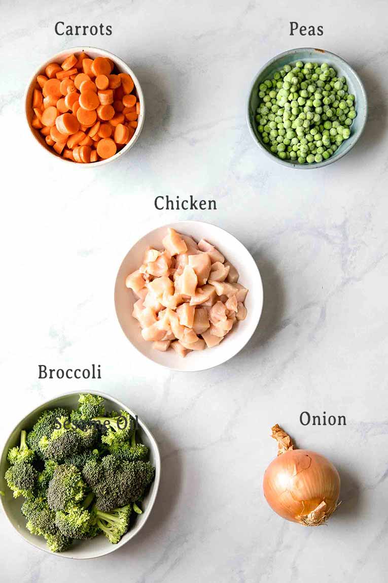Carrots, peas, chicken, broccoli, and onion on a white marble board.