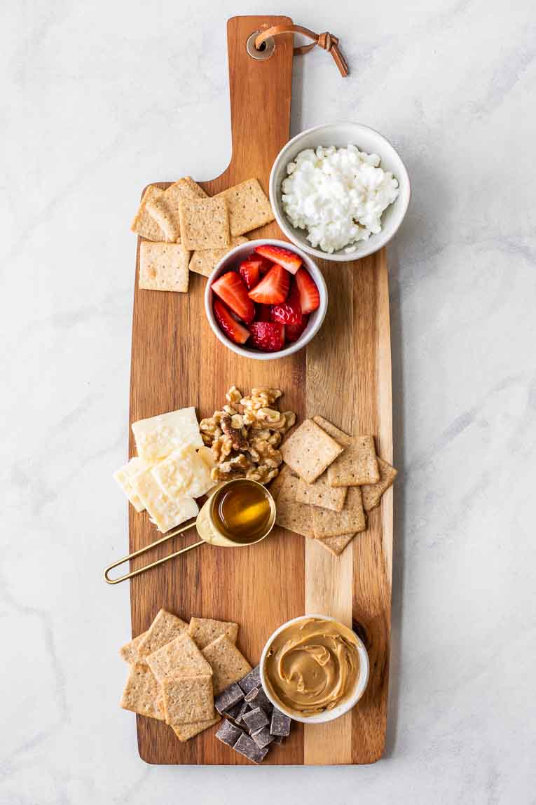 Wooden cheese board with crackers, cheese, fruit, and peanut butter.