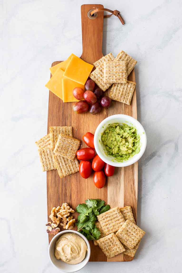 Cheese board with guacamole, crackers, tomatoes, cilantro, cheese, and grapes.