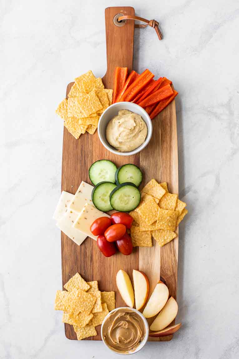 Cheese board with apples, peanut butter, crackers, cucumbers, red bell peppers, and tomatoes.