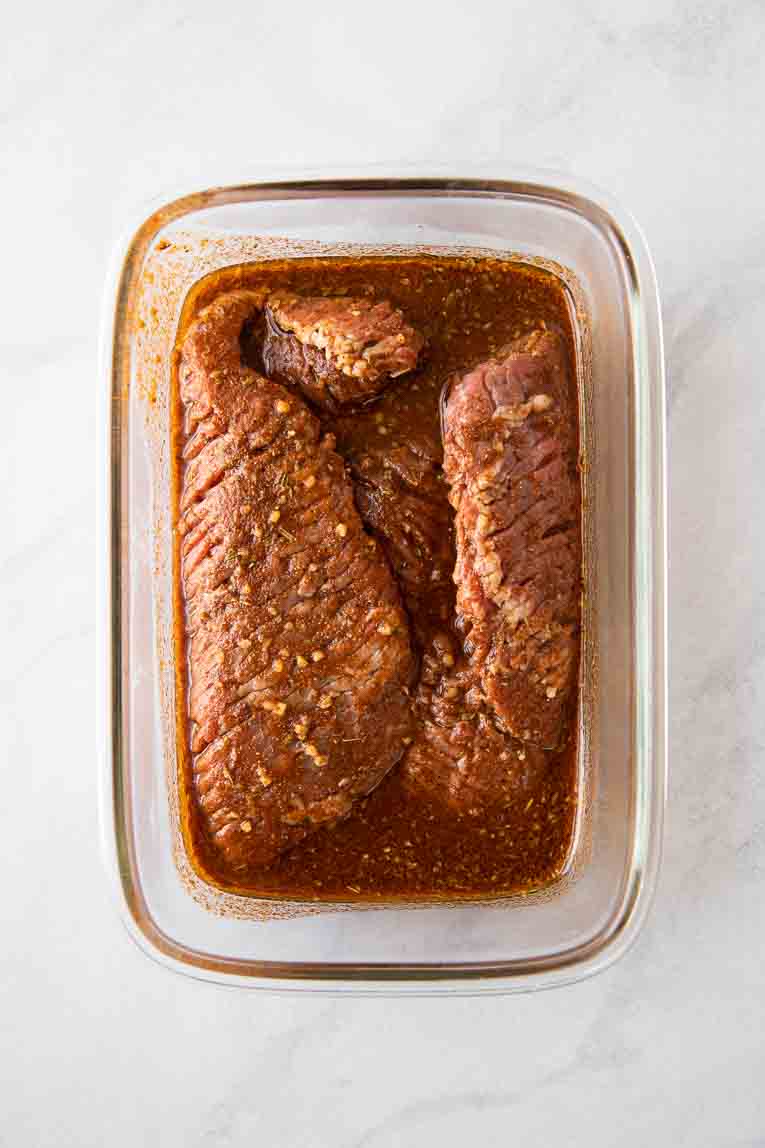 Flank steak being marinated in a glass container.
