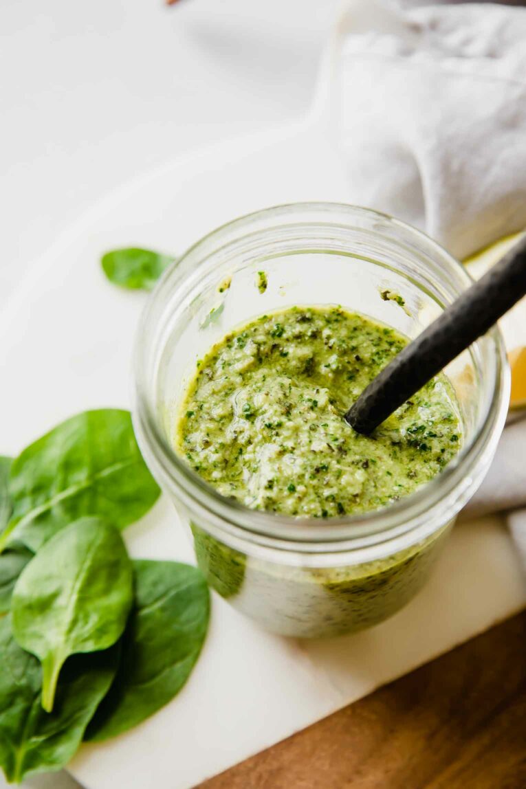 A jar of pesto spinach sauce with a spoon and spinach leaves around the edges. 