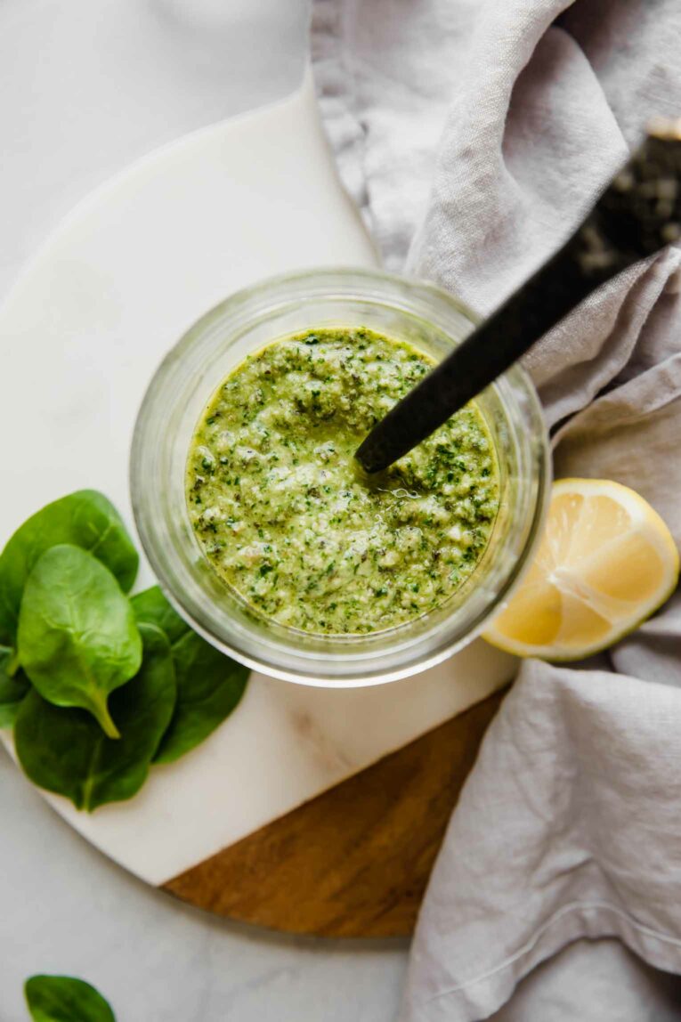 A jar of pesto sauce with spinach leaves and a lemon wedge around it.