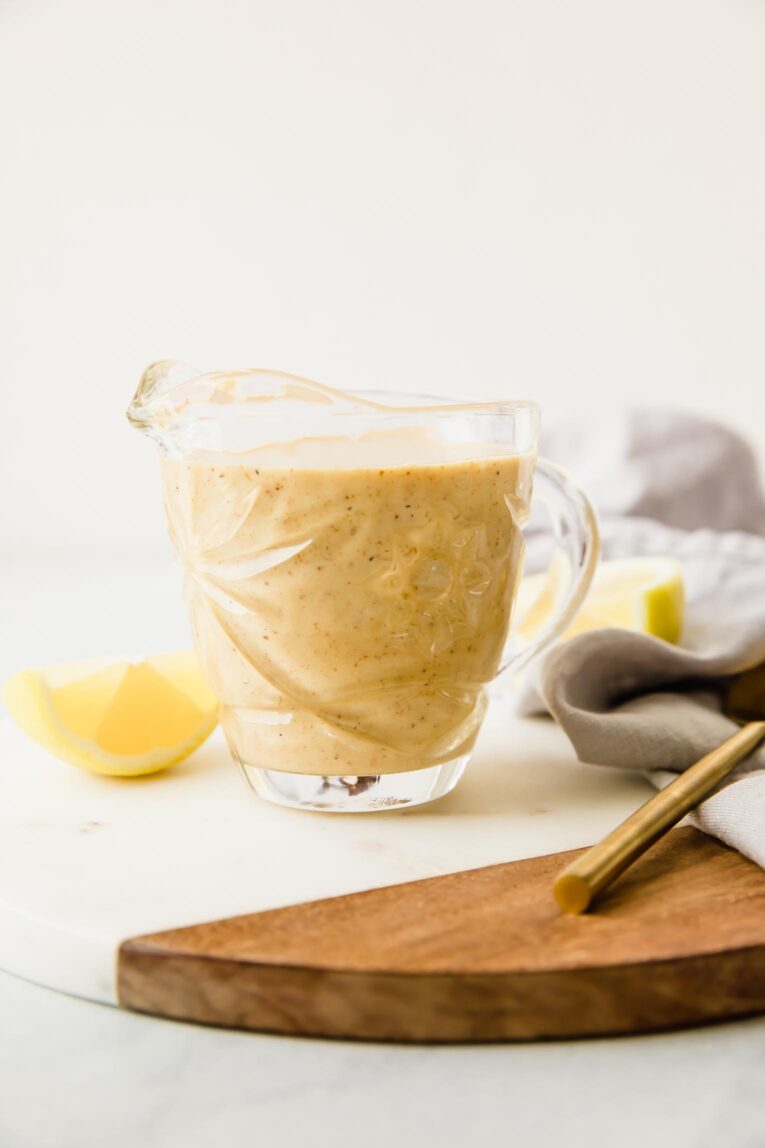 A glass creamer with honey mustard dressing inside sitting on a wooden cutting board.