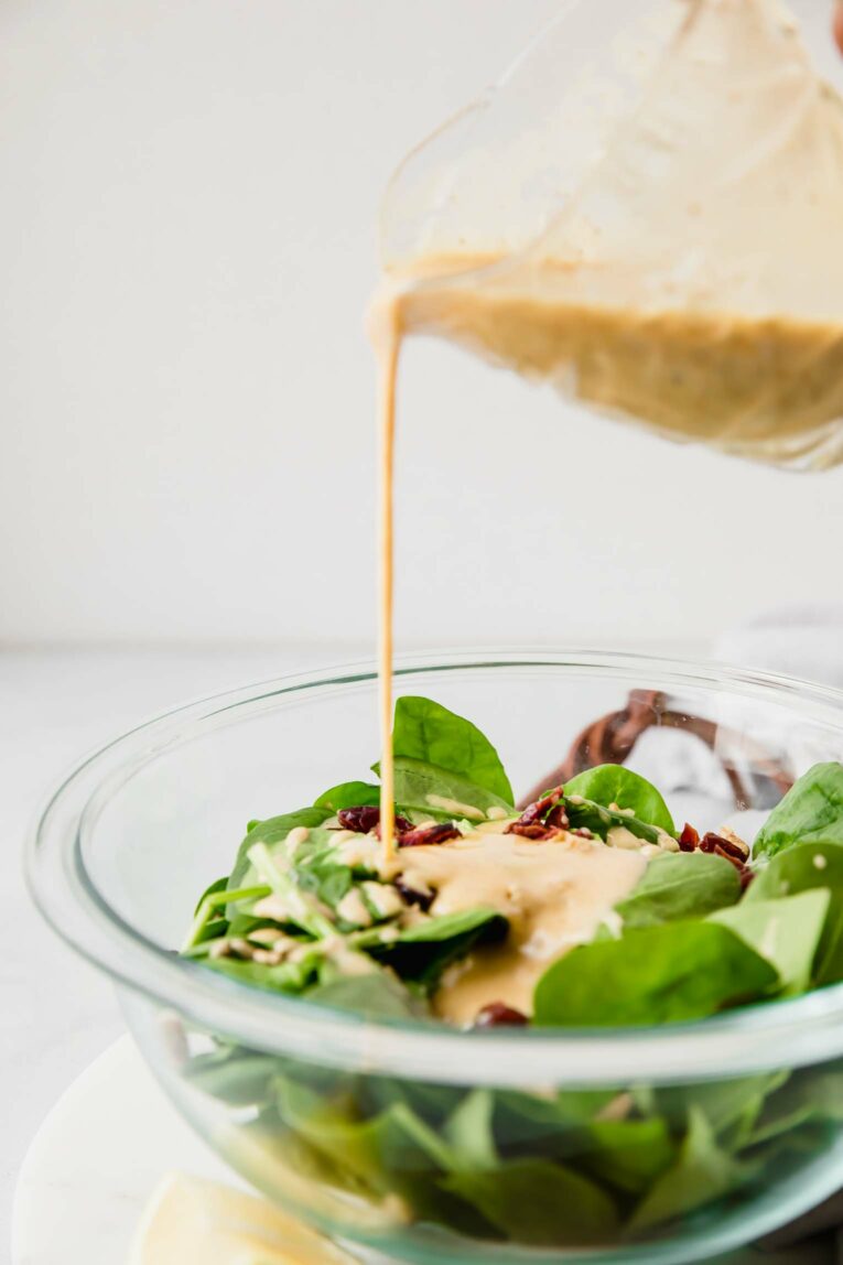 A creamer pouring honey mustard dressing onto a bowl of salad greens.