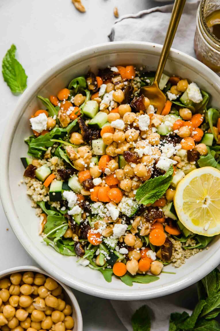 A top shot of a large bowl of carrot chickpea salad with feta cheese and a slice of lemon and a gold fork going into the salad.