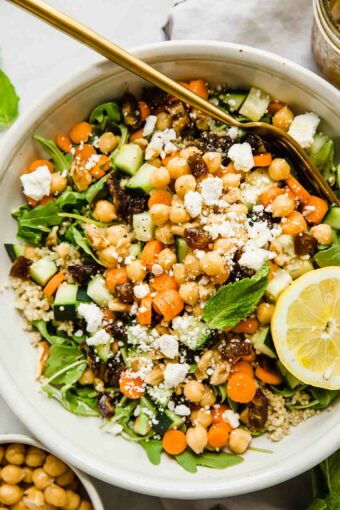 A top shot of a large bowl of carrot chickpea salad with feta cheese and a slice of lemon with a gold fork.