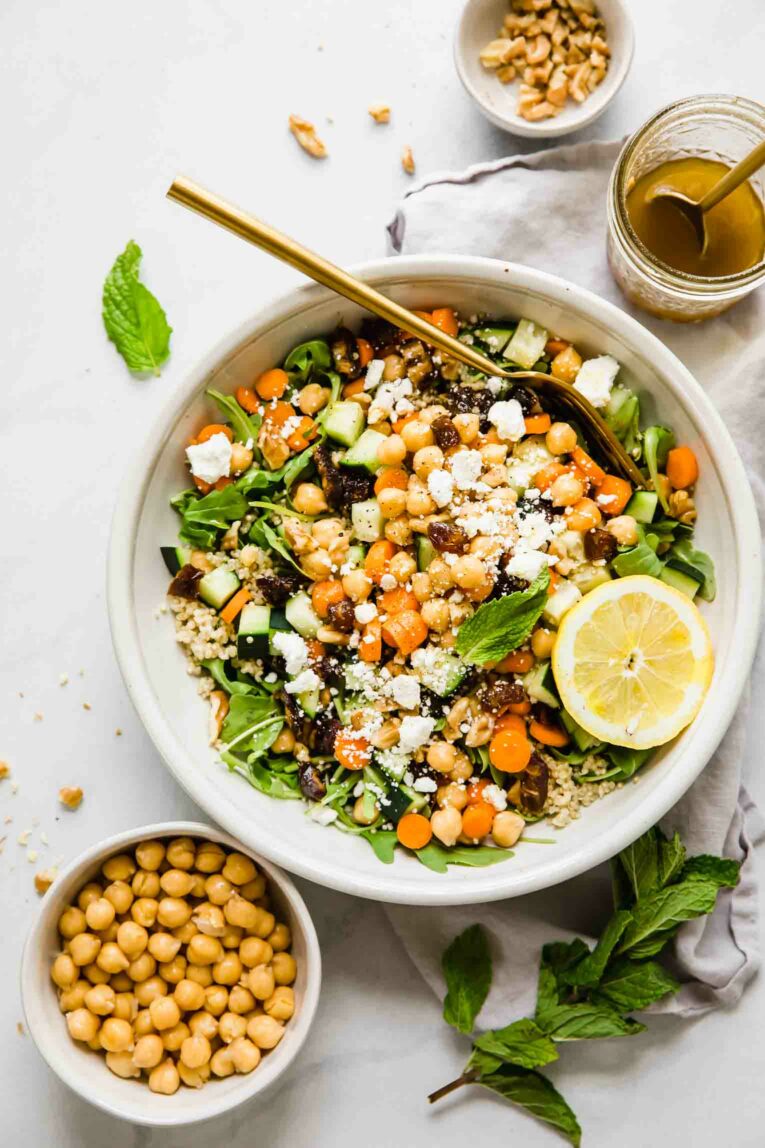A top shot of a large bowl of carrot chickpea salad with feta cheese and a slice of lemon with a cup of dressing on the side.