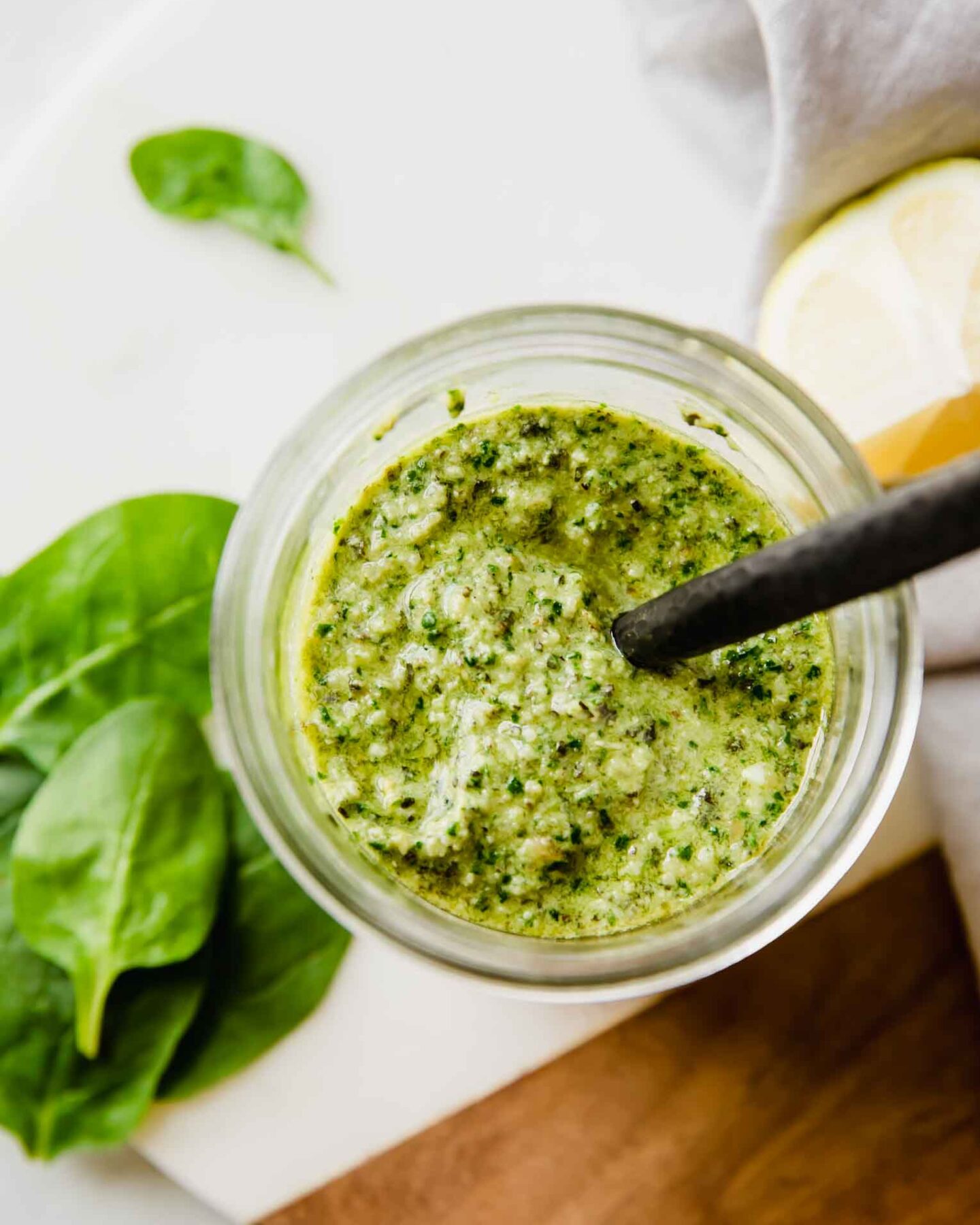 A jar of pesto pasta with a spoon on top of a cutting board and basil leaves around it.