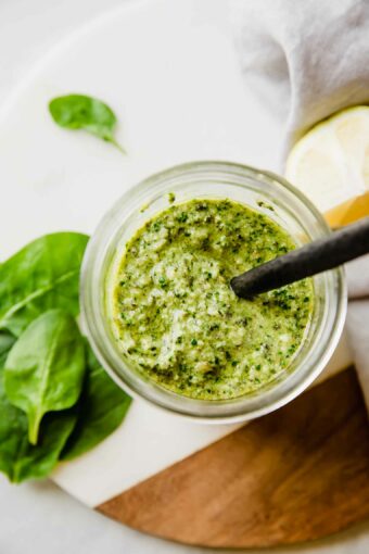 A jar of pesto pasta with a spoon on top of a cutting board and basil leaves around it.