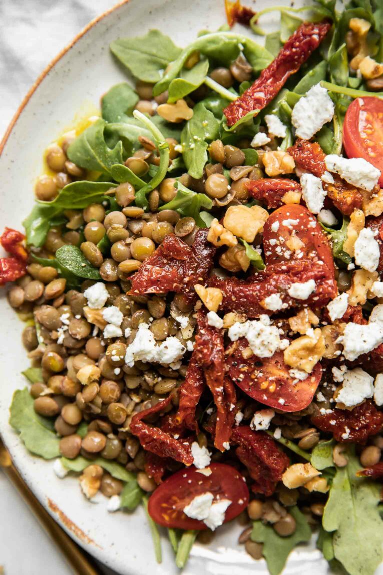 Close up of arugula, lentils, sun-dried tomatoes, walnuts, and goat cheese on a white plate.