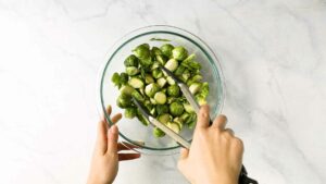 Hands tossing Brussels sprouts in seasoning and oil in a mixing bowl.