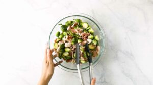 Hands tossing seasoned Brussels Sprouts with bacon in a mixing bowl.