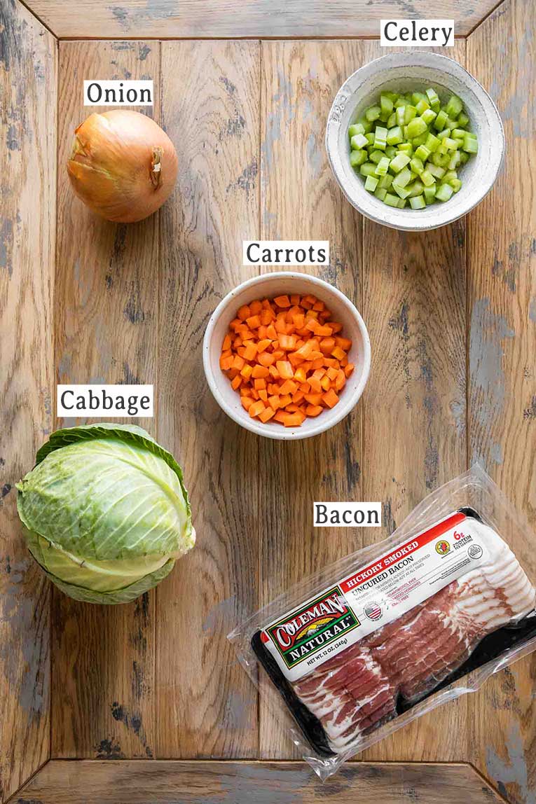 Onion, celery, carrots, cabbage, and bacon on a table.