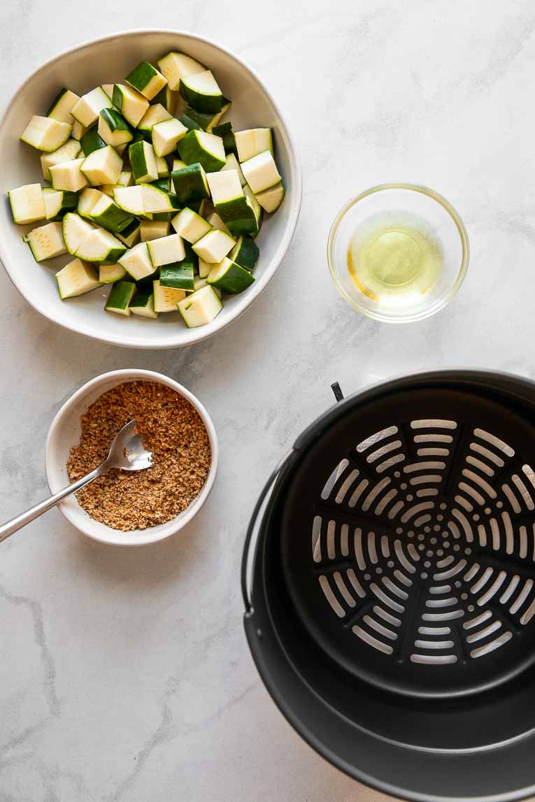Zucchini, seasoning, oil, and air fryer basket on a countertop.