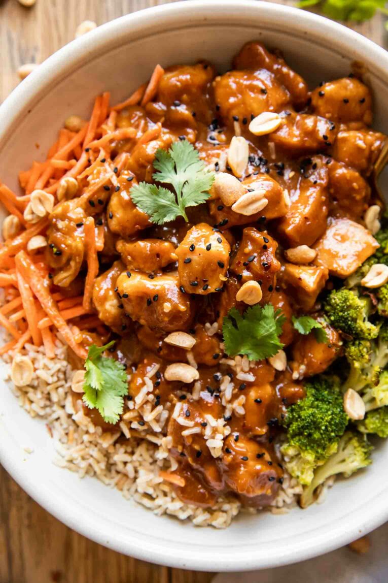 Peanut butter chicken in a bowl with rice, carrots, broccoli, cilantro, and sesame seeds.