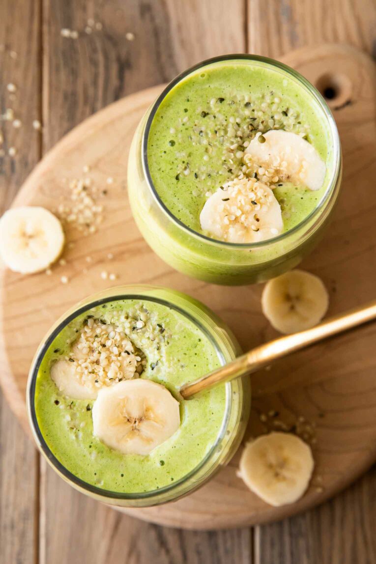 Banana and spinach smoothie in glasses with straws and sliced bananas on top.