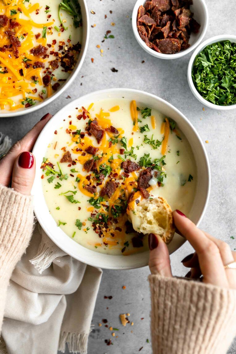Hands dipping bread into a bowl of creamy potato soup.