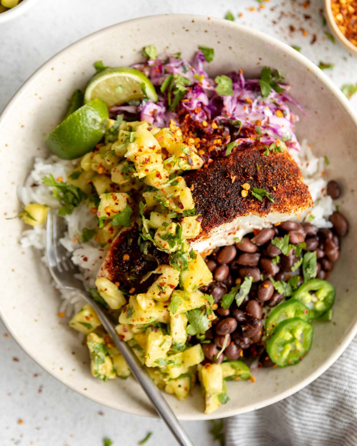 Close up of mahi mahi fish, pineapple avocado salsa, cilantro, lime, jalapeño, and beans and rice in a bowl.