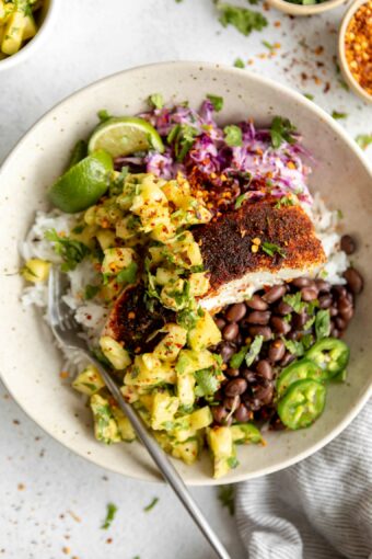 Close up of mahi mahi fish, pineapple avocado salsa, cilantro, lime, jalapeño, and beans and rice in a bowl.