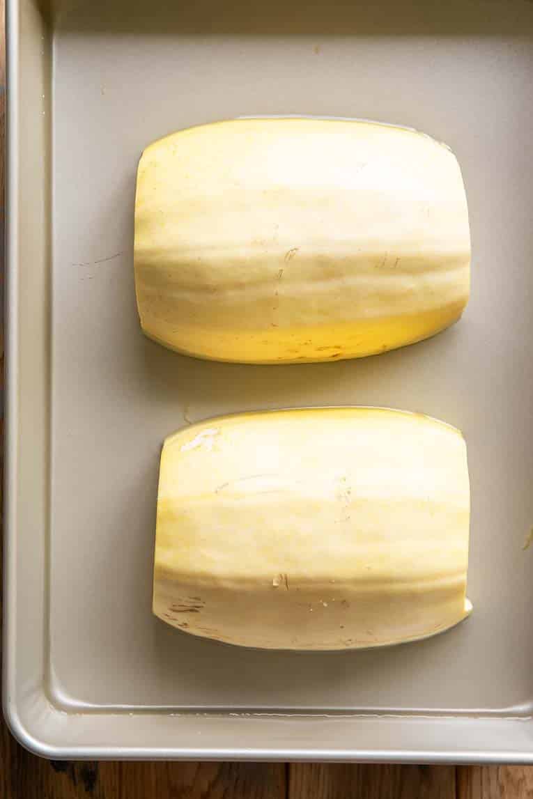 Two spaghetti squash halves upside down on a baking sheet with water.